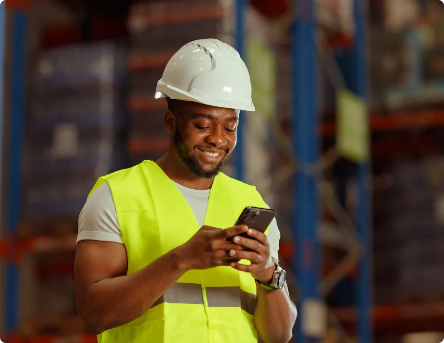 A worker in a hard hat and safety vest smiles while using a smartphone in a warehouse