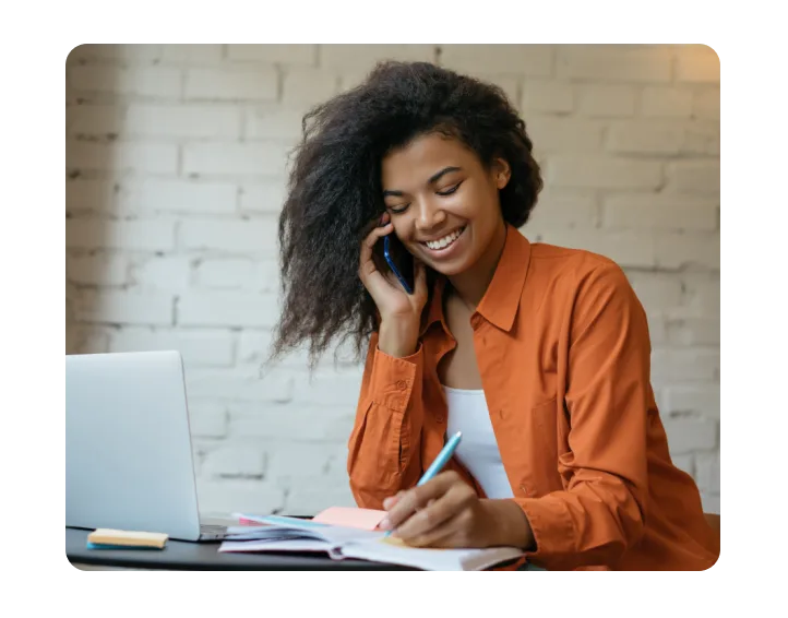 Femme portant une chemise orange, prenant des notes à la main au téléphone, assise devant un ordinateur portable ouvert.