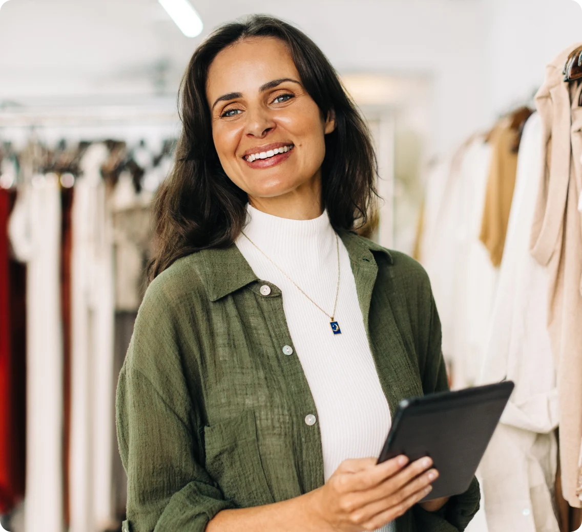 Retail owner smiles, holding a tablet among clothing racks.