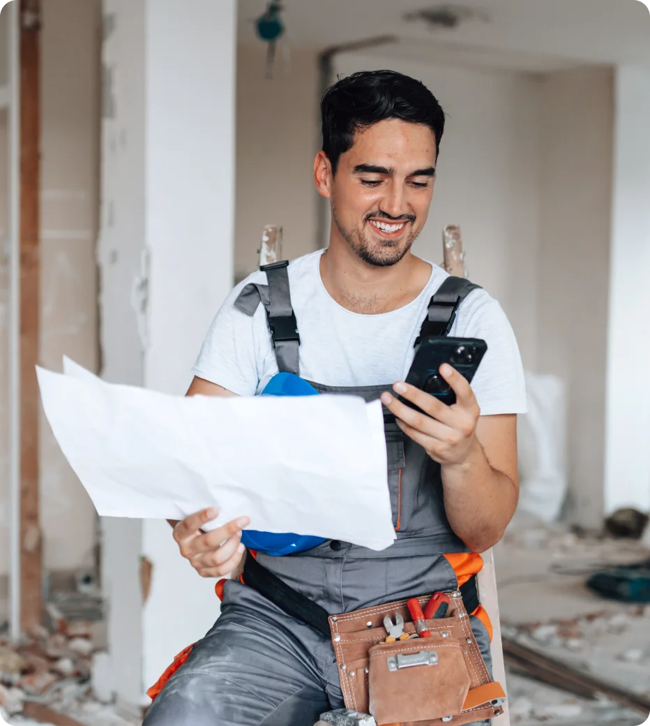 Construction worker in overalls smiling while holding blueprints and a smartphone inside a partially built interior.