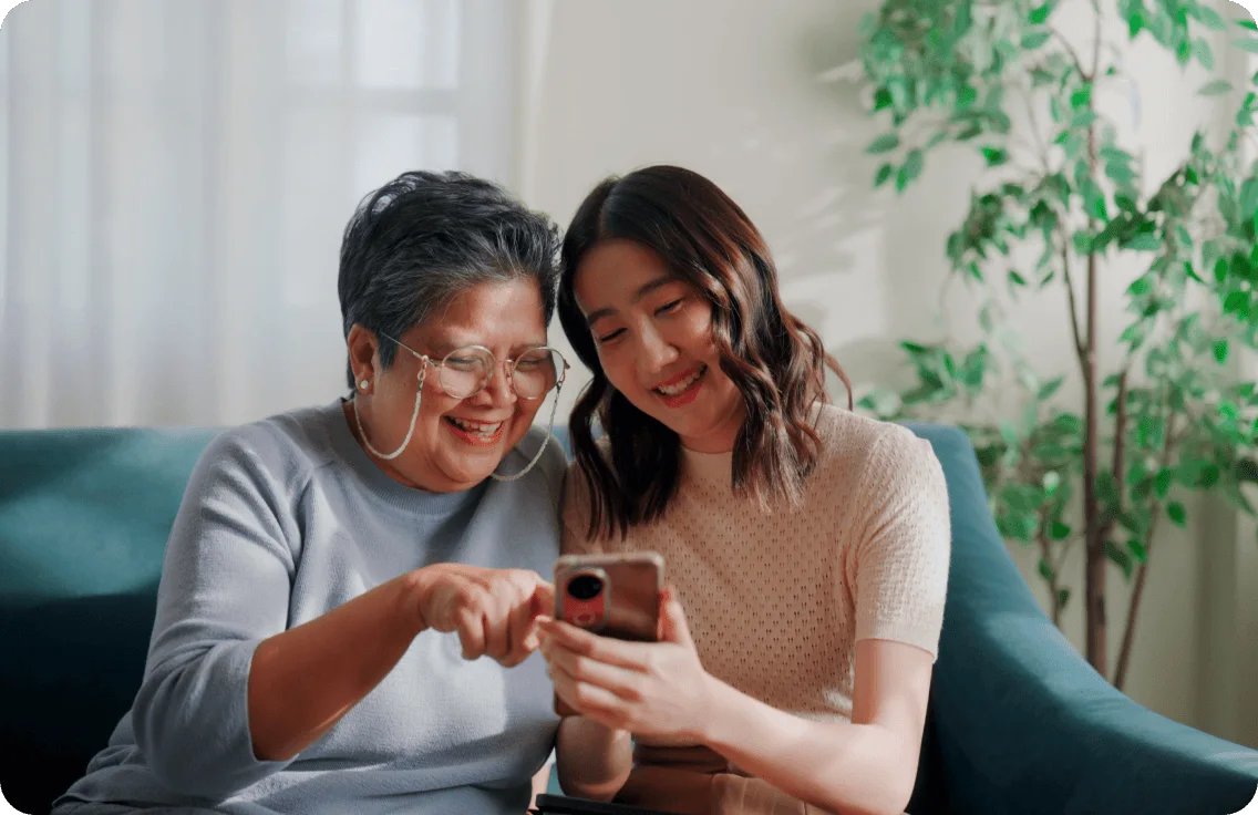 Two women sit smiling on a couch in a sunlit room as the younger woman helps guide the older woman on how to use her new smartphone.
