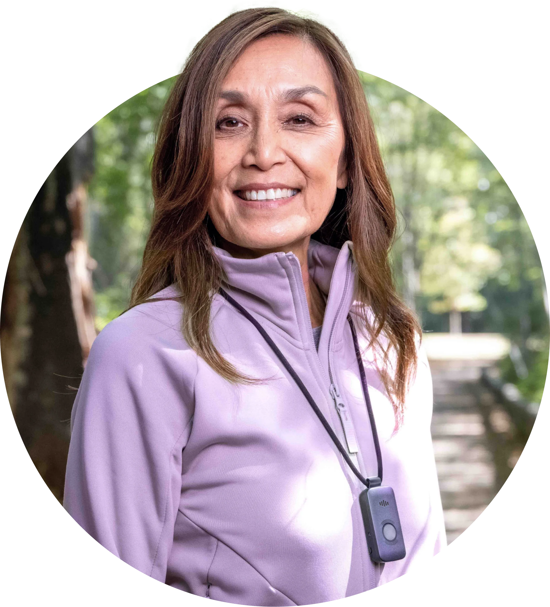A smiling woman standing in a sunny trail wearing a a TELUS Health Medical Alert Pendant