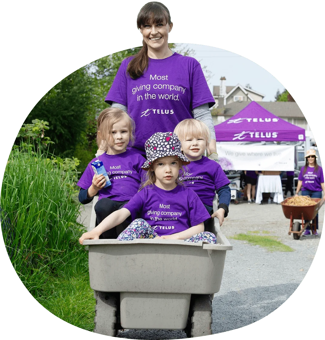 A TELUS volunteer standing behind three children seated in a wheelbarrow