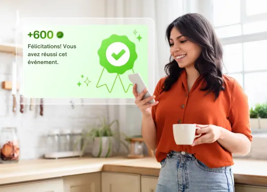 Une personne tient un téléphone intelligent et une tasse à café dans une cuisine, avec un message instantané à l’écran disant : « Félicitations! Vous avez réussi cet événement » et « Plus de 600 points ».