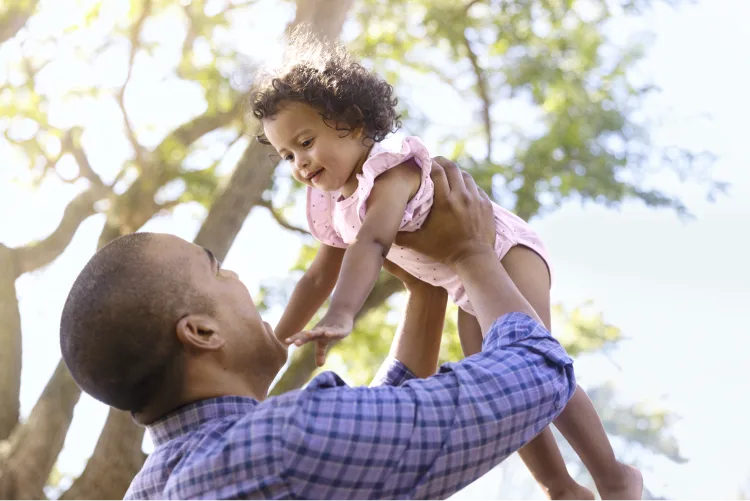 A man lifting up a young child into the air, with sunlit trees in the background.