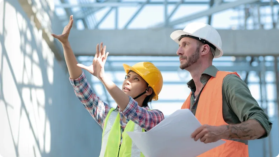 Two people dressed in hard hats and safety vests are reviewing building plans and assessing progress on the building they are inspecting.