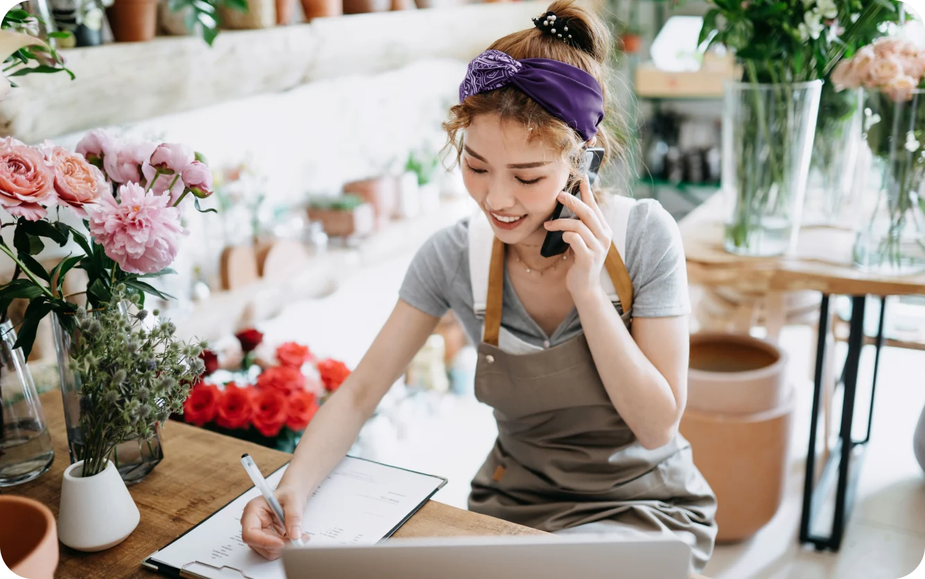 Un fleuriste souriant en tablier gris parle au téléphone et écrit sur une planchette à pince. La boutique est remplie de plantes et de fleurs en pot.