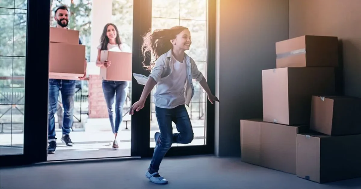 A young girl runs excitedly into her new home followed by her parents holding cardboard moving boxes.