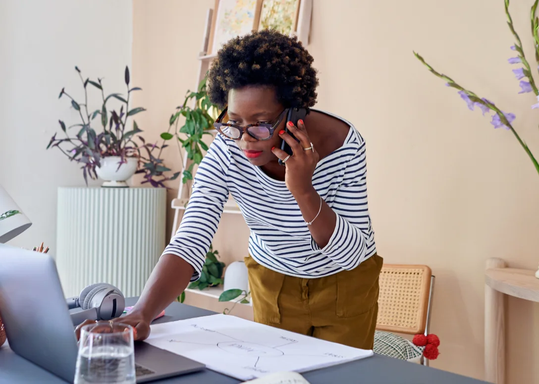 Woman stands, her brows furrowed, hovering over an open laptop, while having a conversation on her smart phone