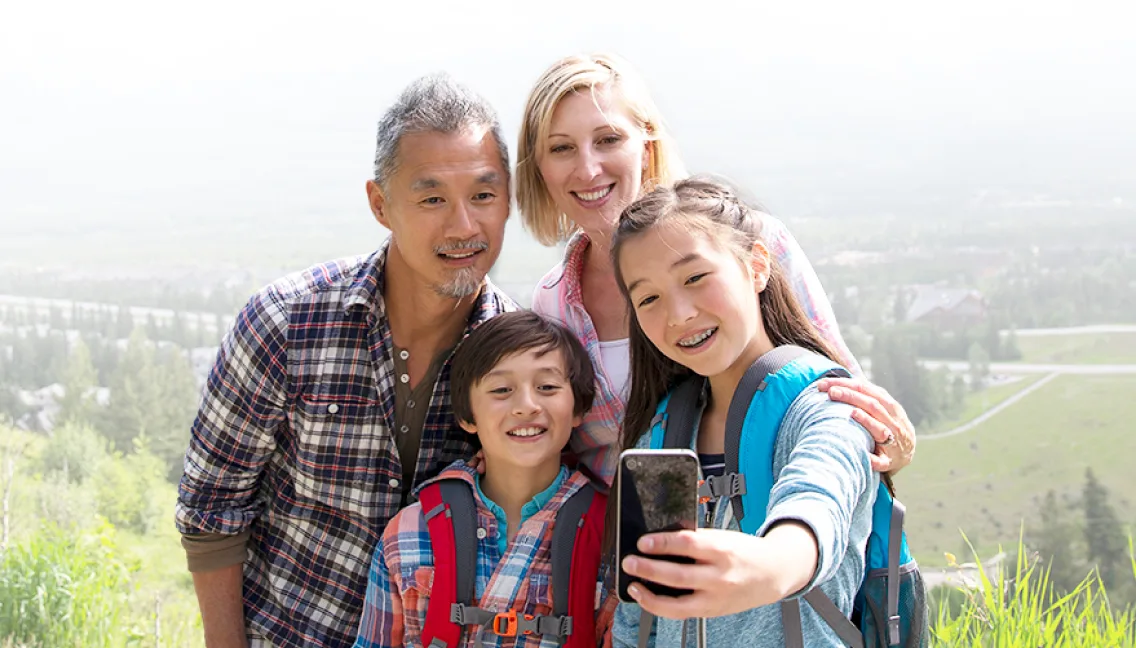 Two parents and two children wearing backpacks pose for a selfie with a lush green valley in the background.