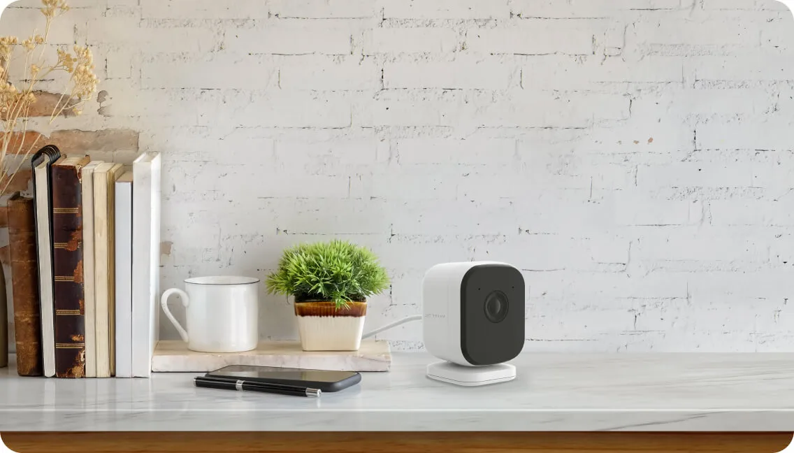 A TELUS Indoor Camera placed on a shelf beside beside a small plant, cup and some books.