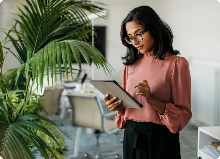 A professional woman with glasses stands in a modern office, looking at and using a digital tablet.