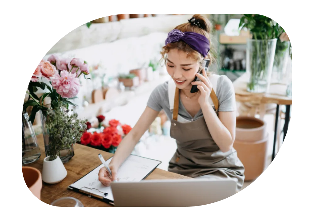 A smiling florist in a gray apron talks on the phone and writes on a clipboard. The shop is filled with potted plants and flowers.