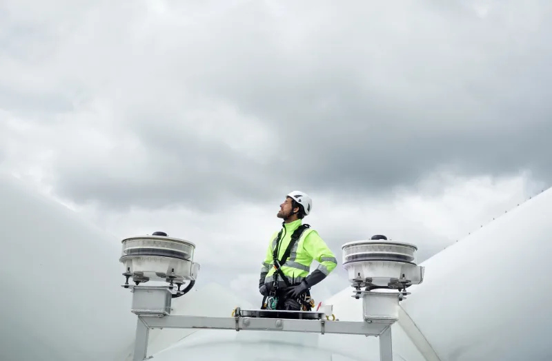 Male worker standing on a wind turbine, wearing a high-visibility safety vest, harness, and hard hat looking off into the distance to the left