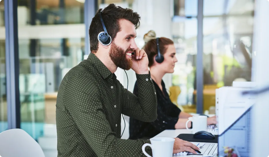 Two colleagues, working as help-desk support, wearing headsets, seated in front of computers