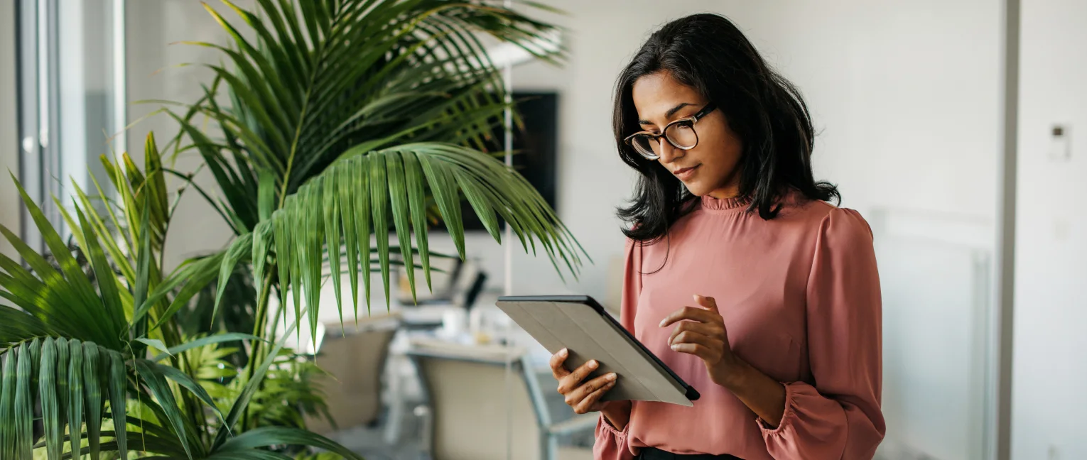 A professional woman with glasses stands in a modern office, looking at and using a digital tablet.