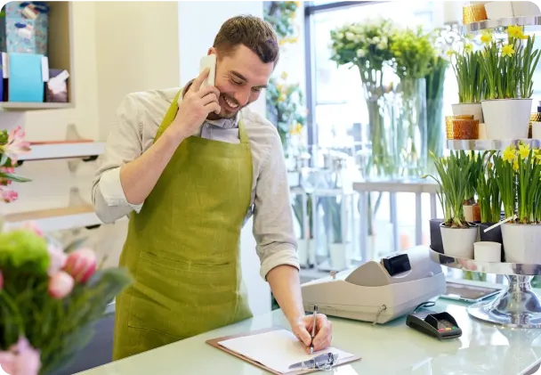 A smiling florist in a green apron talks on the phone and writes on a clipboard. The shop is filled with potted plants and flowers, with a cash register and card reader on the counter, in a bright and inviting space.