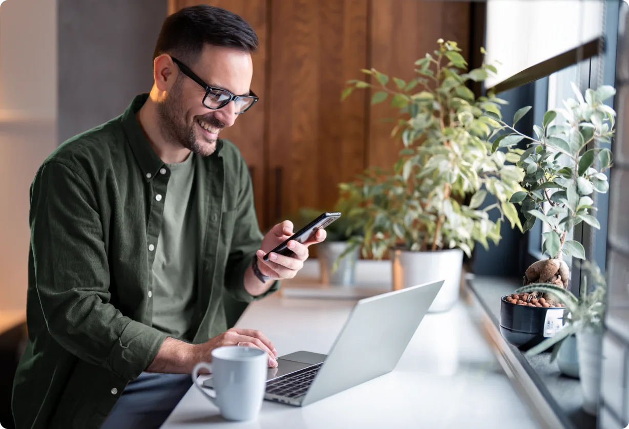 Man checks his phone beside a laptop in a bright office.