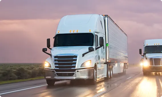White semi-trucks driving on highway at dusk with headlights on.