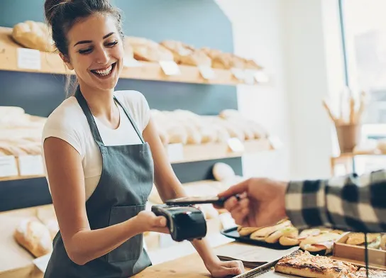 Smiling woman wearing green apron, holding payment machine, working in a bakery. Customer with outstretched hand is using smartphone to pay on the payment machine.