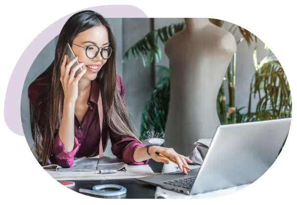 Femme en conversation au téléphone, assise devant un ordinateur portable ouvert. Une forme de robe est aussi visible.