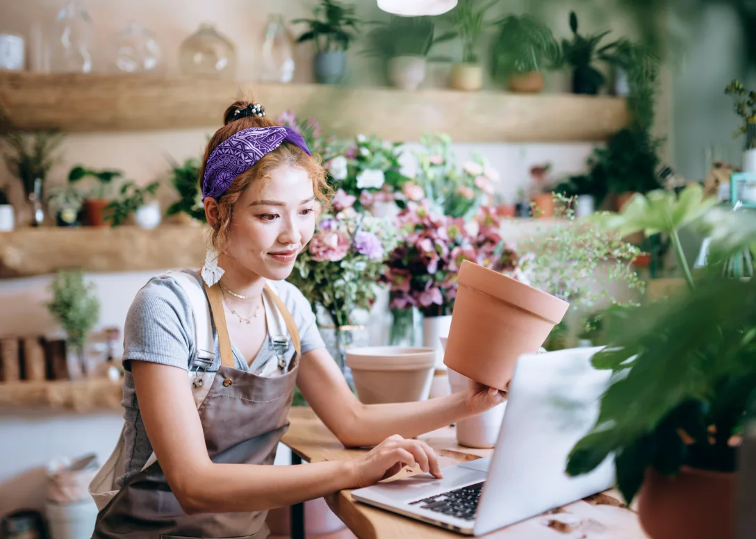 Person using a laptop while holding a flower pot in a flower shop.