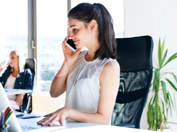 Une femme assise sur une chaise est au téléphone et travaille à l’ordinateur.