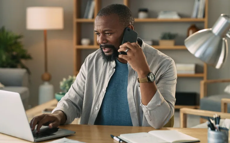 A smiling businessman sitting at a desk in front of a laptop talking on a cell phone