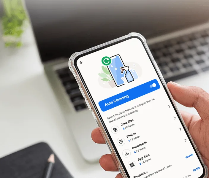 A hand holding a smartphone displaying the Norton Utilities Ultimate app with the "Auto Cleaning" feature enabled. A laptop is blurred in the background.