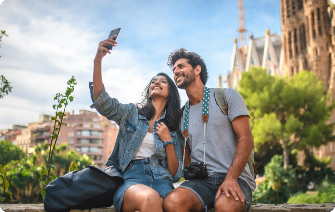 A happy couple sitting outdoors on vacation, smiling as they take a selfie together with a smartphone.