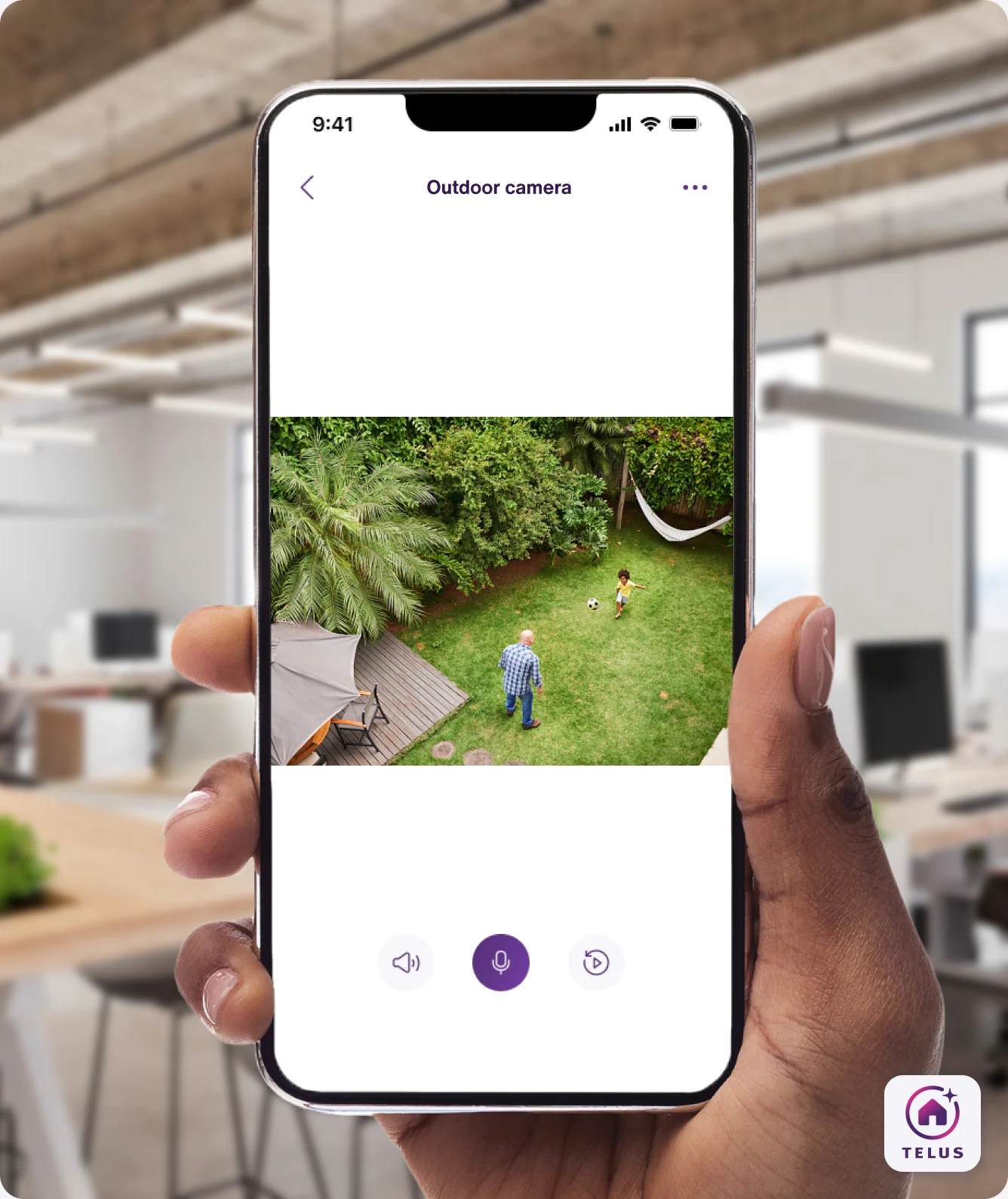 A hand holding the TELUS SmartHome+ app showing a family playing in the backyard from the TELUS Outdoor Camera. 