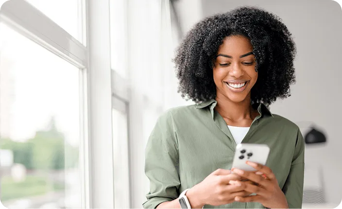 A smiling woman looking at her smartphone next to a bright window.