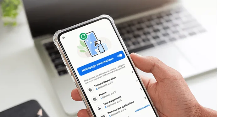 Une main tenant un téléphone intelligent qui affiche l’application Norton Utilities Ultimate avec la fonction de nettoyage automatique activée. Un ordinateur portable est flou en arrière-plan.