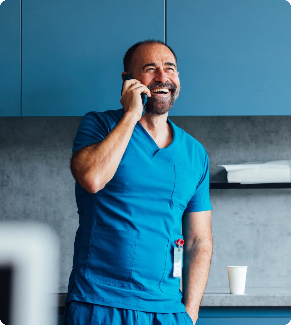 Smiling male healthcare worker in blue scrubs on a mobile call near a clinic setting.