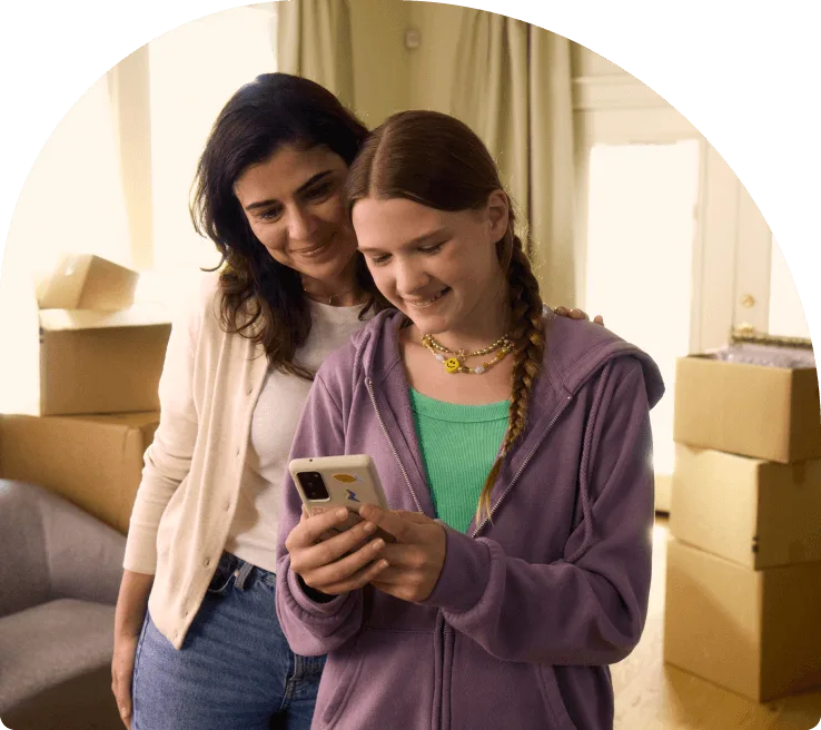 A mother and daughter standing in their living room and viewing a smartphone together.