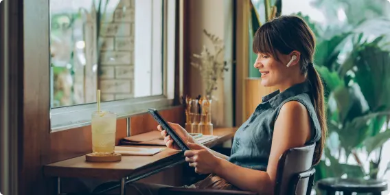 Une femme souriante est assise à un bureau. Elle porte des écouteurs et regarde une tablette.