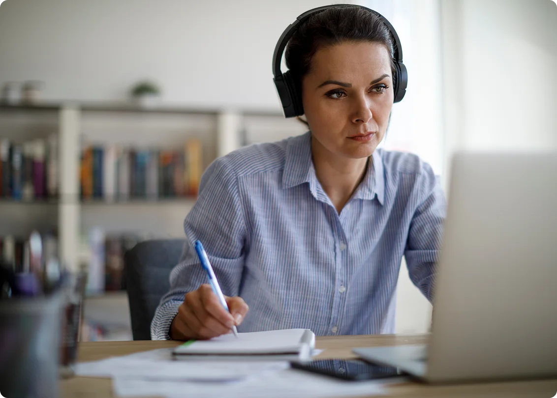 Professional with headset taking notes during virtual meeting at home office.