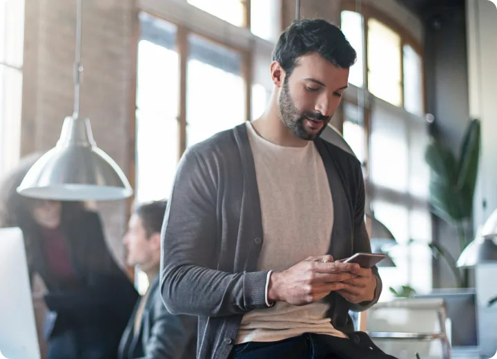 Man in a bright office checks his smartphone while seated on a desk.