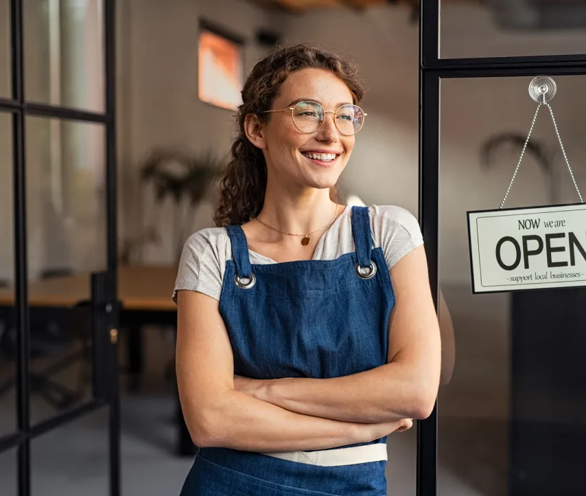 Une femme souriante se tient fièrement devant sa boutique qui vient d’ouvrir.