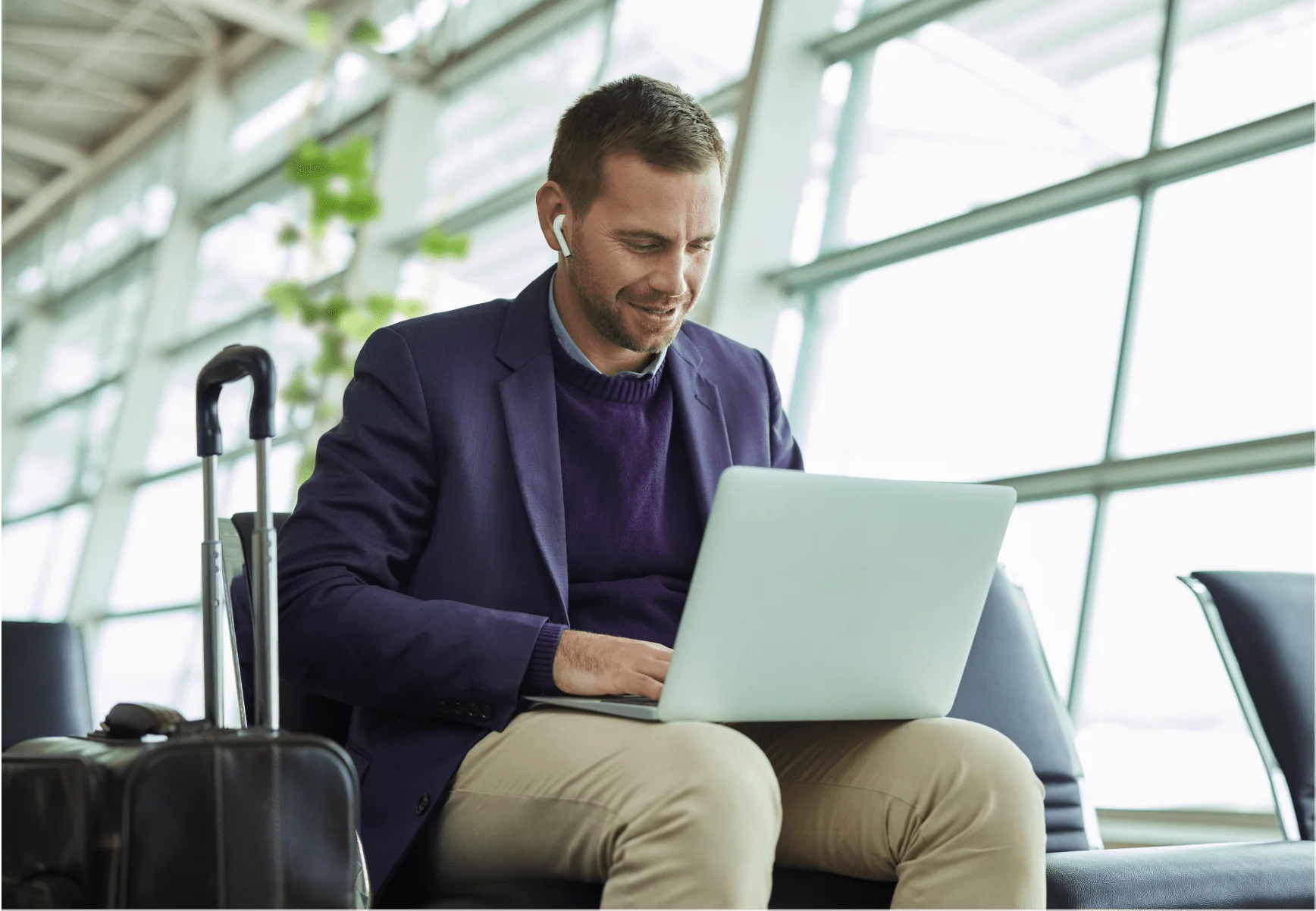 Un homme bien habillé assis dans une zone d'attente d'un aéroport – il porte des écouteurs et s’affaire sur son ordinateur portable.