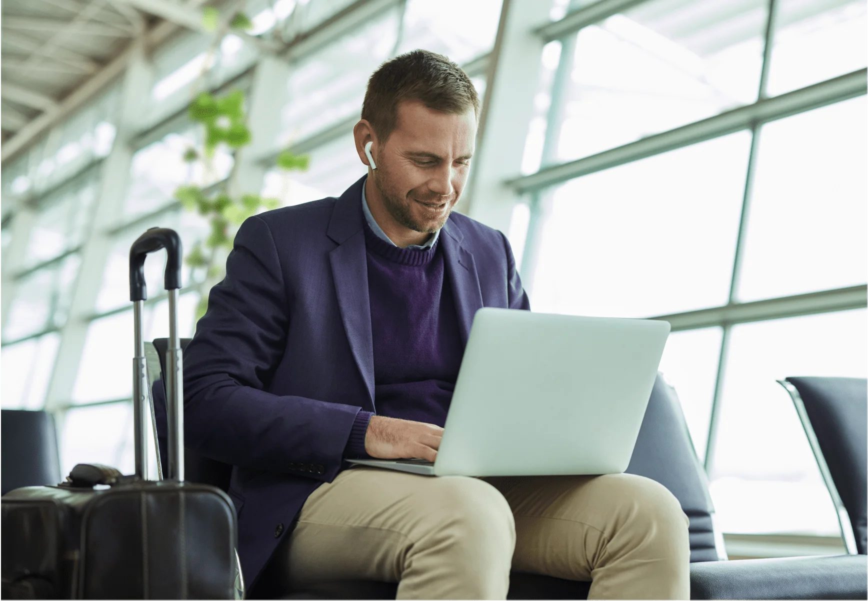 A well-dressed man sitting in an airport waiting area - he is wearing earbuds and doing something on his laptop.