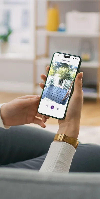 Une personne tient un téléphone intelligent affichant une vidéo en direct provenant d'une sonnette vidéo, montrant un porche et de la verdure.
