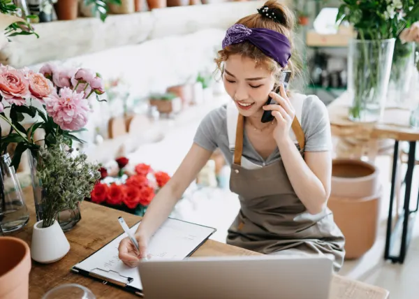 A smiling florist in a gray apron talks on the phone and writes on a clipboard. The shop is filled with potted plants and flowers.