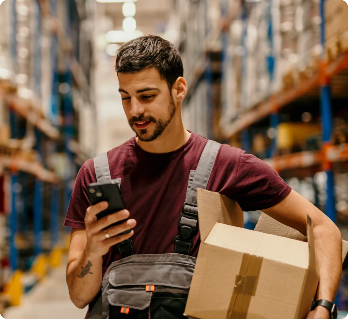 A warehouse worker carries a box and checks his smartphone between aisles.
