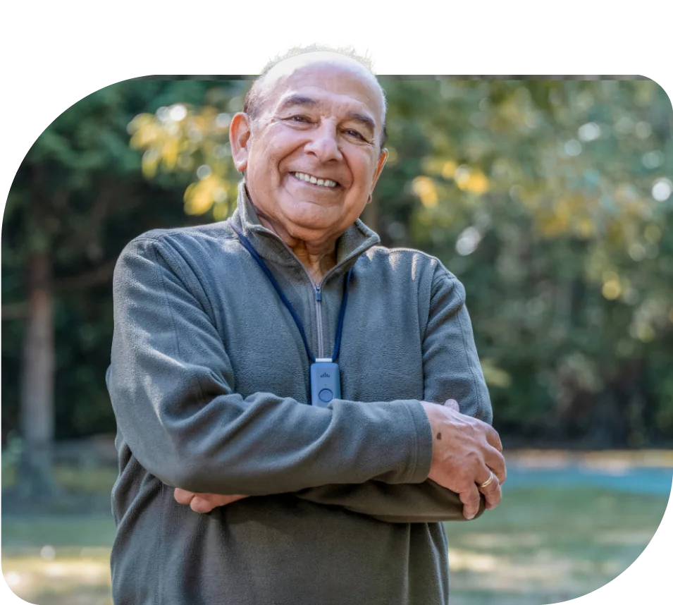 A older man smiling in a park wearing a TELUS Health Medical Alert Pendant
