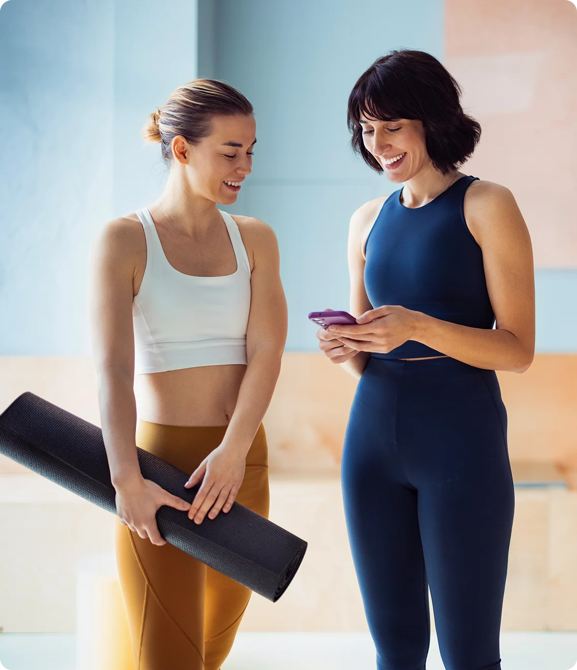 Two women in athletic wear smile at a phone; one holds a yoga mat.