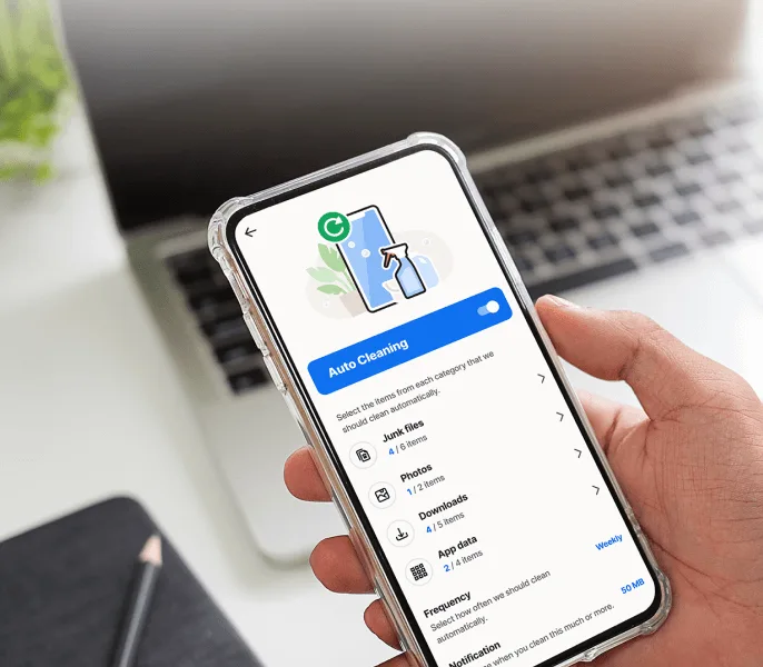 A hand holding a smartphone displaying the Norton Utilities Ultimate app with the "Auto Cleaning" feature enabled. A laptop is blurred in the background.