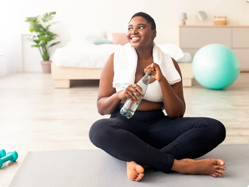 Woman sitting on exercise mat at home holding a water bottle, with dumbbells and fitness equipment nearby.