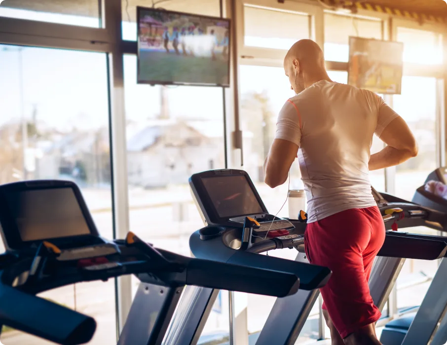 Man runs on a treadmill in a gym with TVs playing overhead.