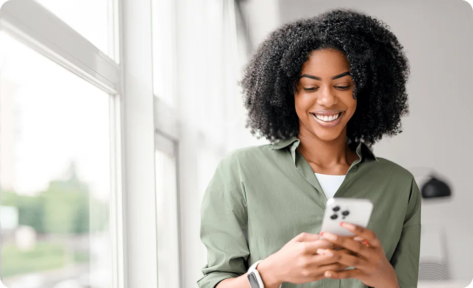 A smiling woman looking at her smartphone next to a bright window.